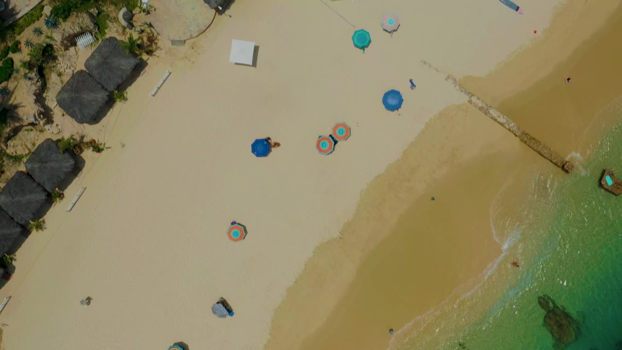 Top-down aerial view of Palmilla Beach with pale sand, palm trees, beach umbrellas, a small shoreline inlet, and tiny distant people that are not recognizable.