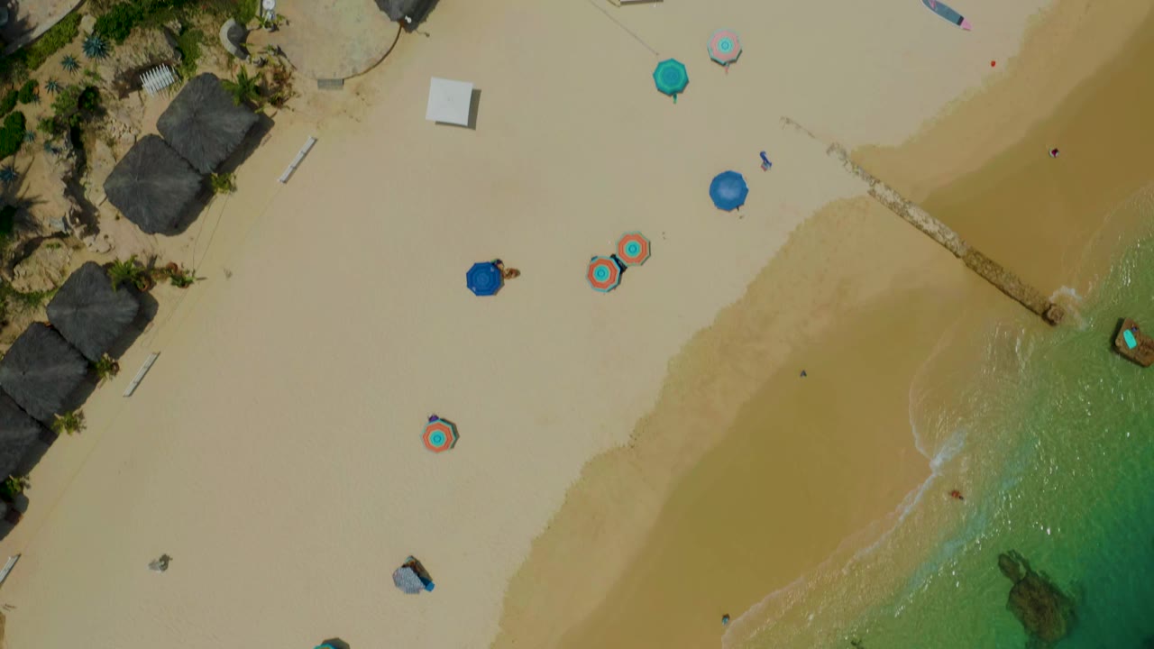Overhead beach view with shoreline, umbrellas, palms, and resort structures.
