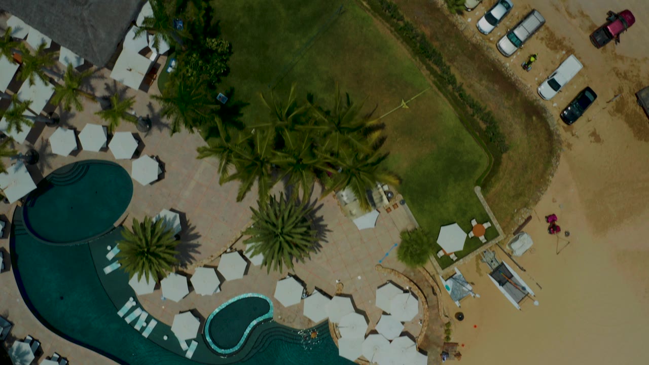Overhead view at Palmilla Beach, Baja California Sur, showing a curved blue pool, white umbrellas, palm trees, green lawn, sandy parking area, and parked cars, with no visible people.