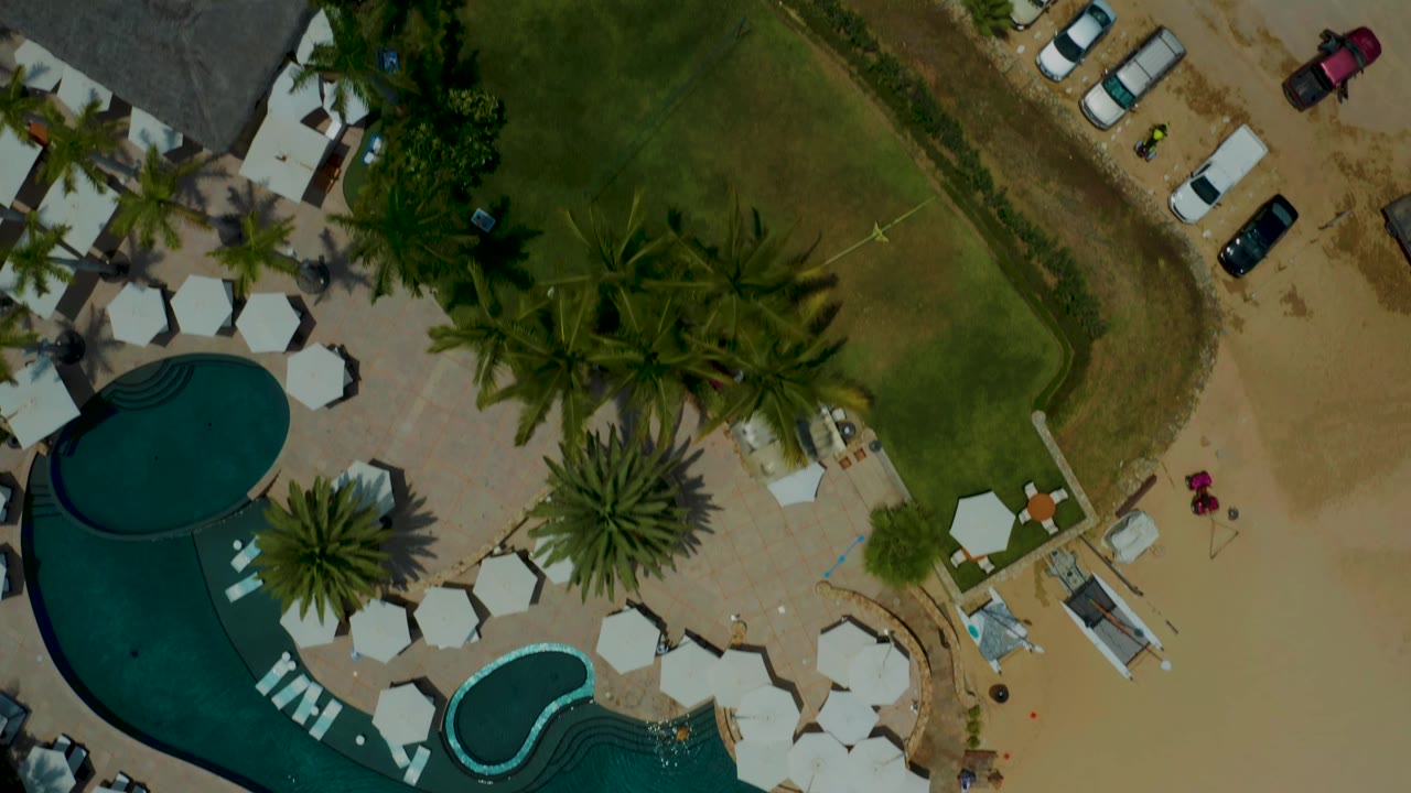 Aerial view of pool, palms, shade umbrellas, grass, sand, and parked cars beside resort buildings.
