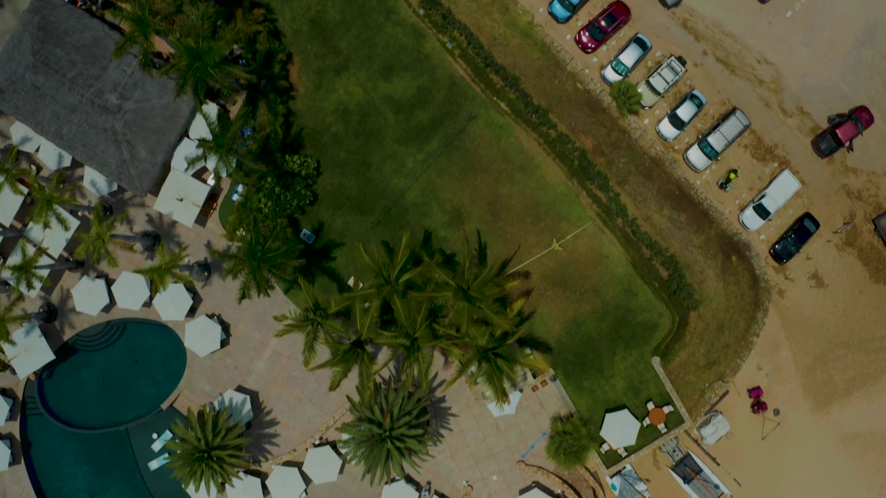 Top-down Palmilla Beach view with the pool partly cropped, palm clusters across the lawn, white umbrellas near resort structures, rows of cars on sand, and no visible people.