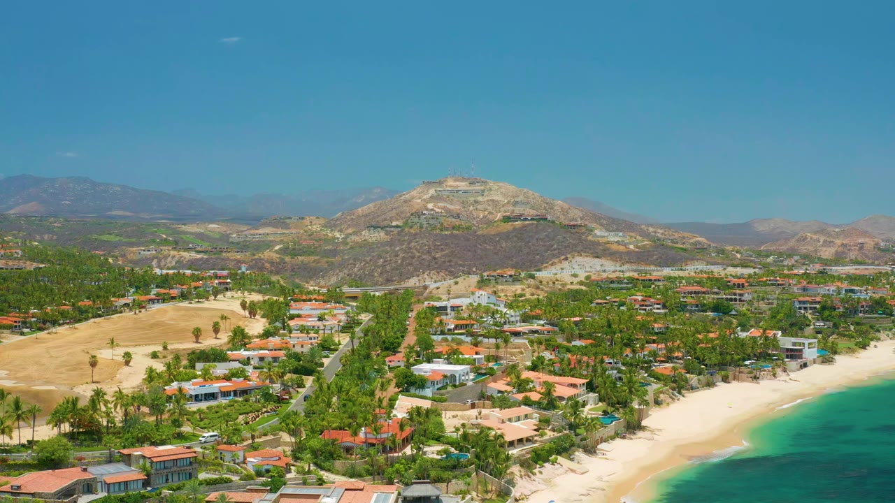 Opening aerial frame over Palmilla Beach, Baja California Sur, with turquoise sea at lower right, pale sand, palm-lined homes, dry hills, distant mountains, and no visible people.