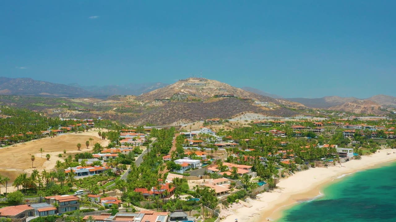 Wide aerial view of Palmilla Beach with the waterline along the lower right, orange-roofed and white homes among palms, dry hills behind, mountains on the horizon, and no visible people.