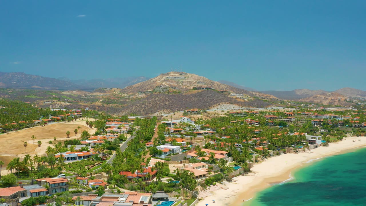 Aerial landscape with sandy beach, bright sea, palms, homes, and mountain ridge.