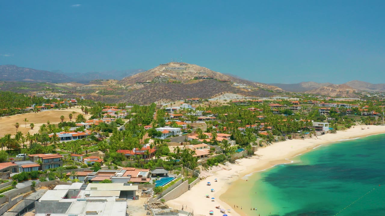 Opening aerial frame over Palmilla Beach, Baja California Sur, with flat-roof homes in the foreground, a curved sand beach, green water, palms, and dry mountains beyond.