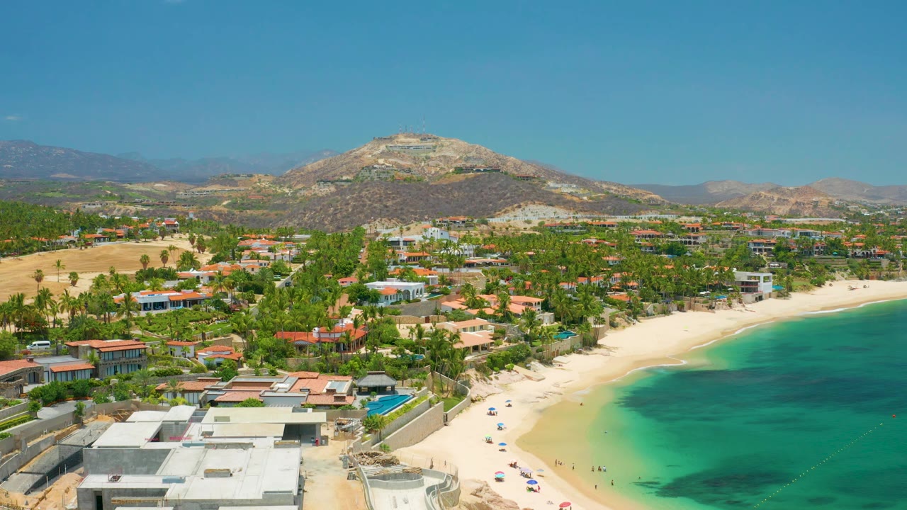 Aerial frame at Palmilla Beach with the bay pushed to the right, beach umbrellas along the sand, clustered palms and houses inland, and a rounded dry mountain under clear sky.