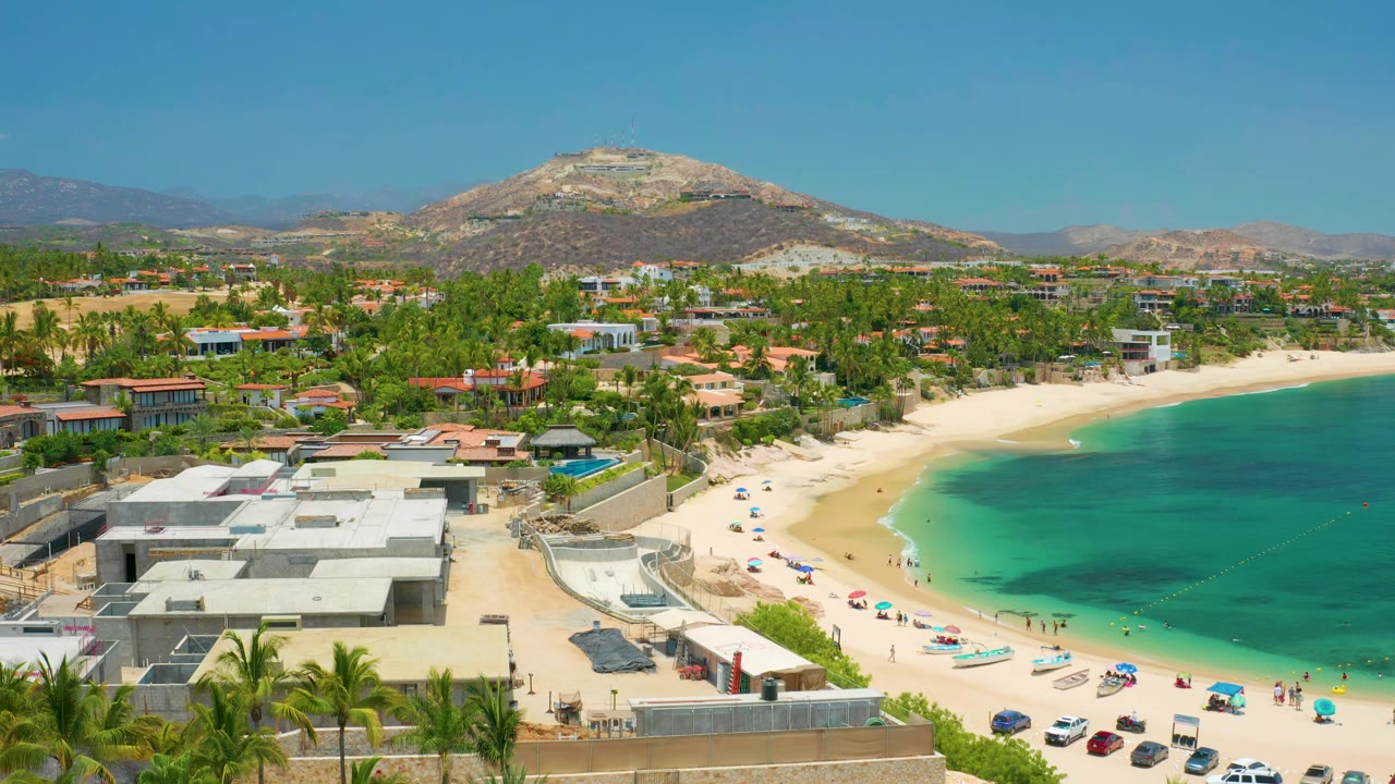 Wide coastal aerial with beachfront buildings, palm trees, bay water, and inland hill.
