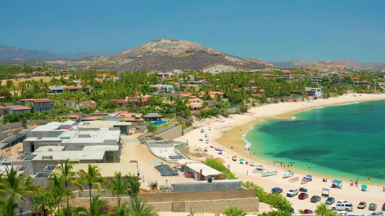 Aerial frame over Palmilla Beach with more of the right-hand bay visible, beach umbrellas and parked cars along the sand, palms at the lower edge, and a rocky hill behind the coastal neighborhood, with no clearly visible people.