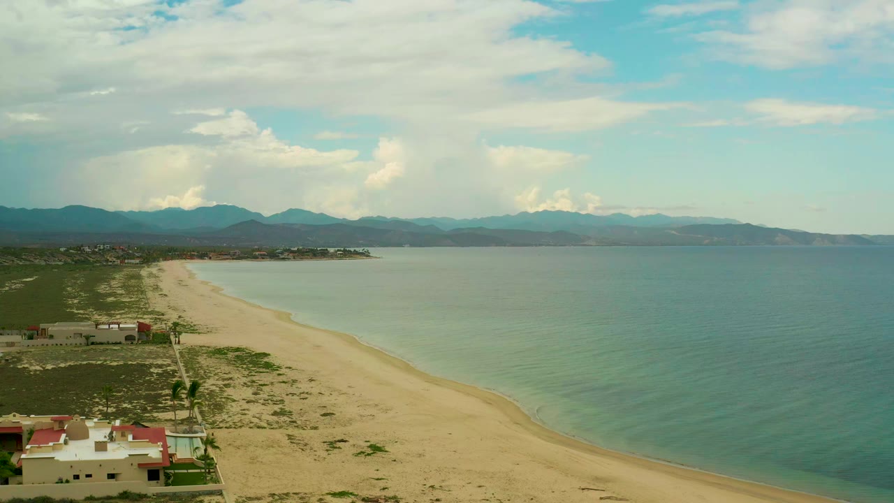 Aerial view over La Ribera, Baja California Sur, with beach-side homes in the lower left, a long sandy shore, blue-green water, distant mountains, clouded sky, and no visible people.