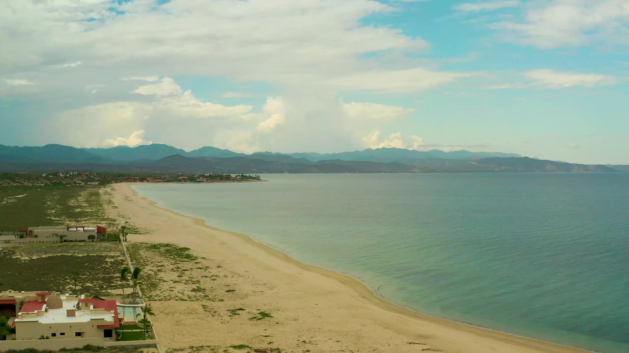 Wide coastal aerial with beach-side houses, pale sand, calm water, and mountains.