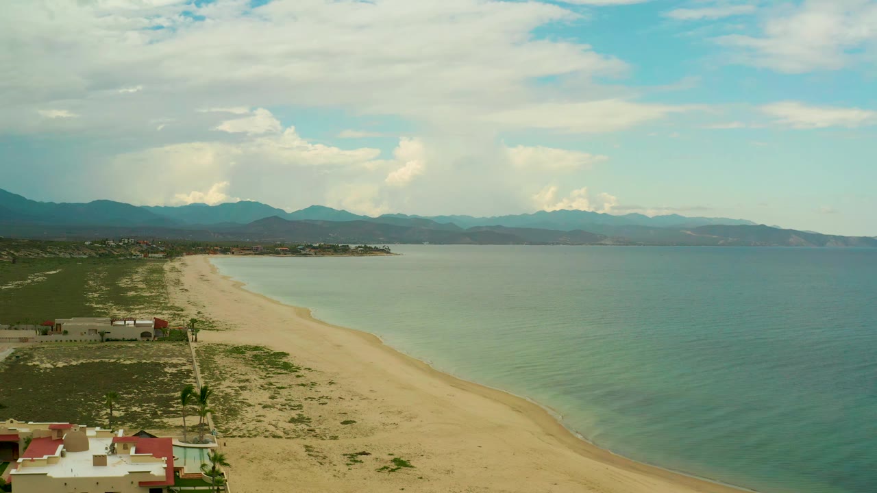 Wide aerial frame over La Ribera, Baja California Sur, showing the beach and waterline slightly farther along the coast, with small buildings at lower left, mountains in the distance, and no visible people.