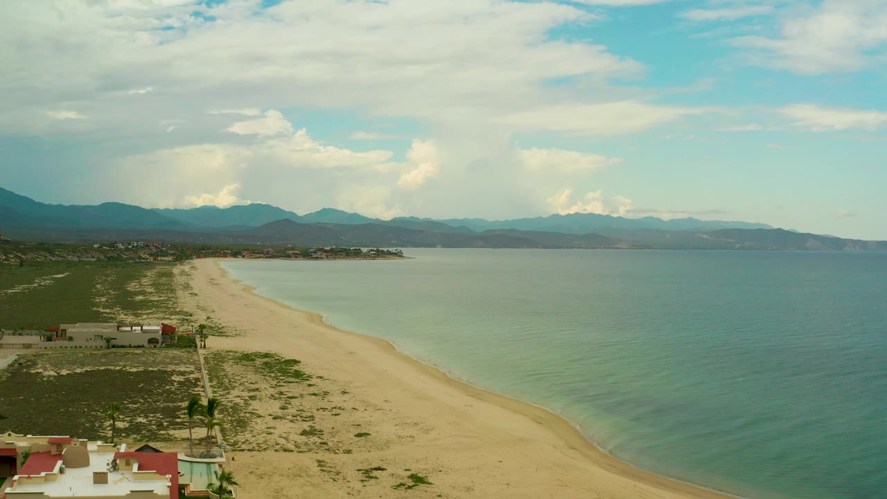 Aerial view of La Ribera, Baja California Sur, with the curved beach leading toward a low point of land, pale water near shore, darker mountain ridges behind, and no visible people.