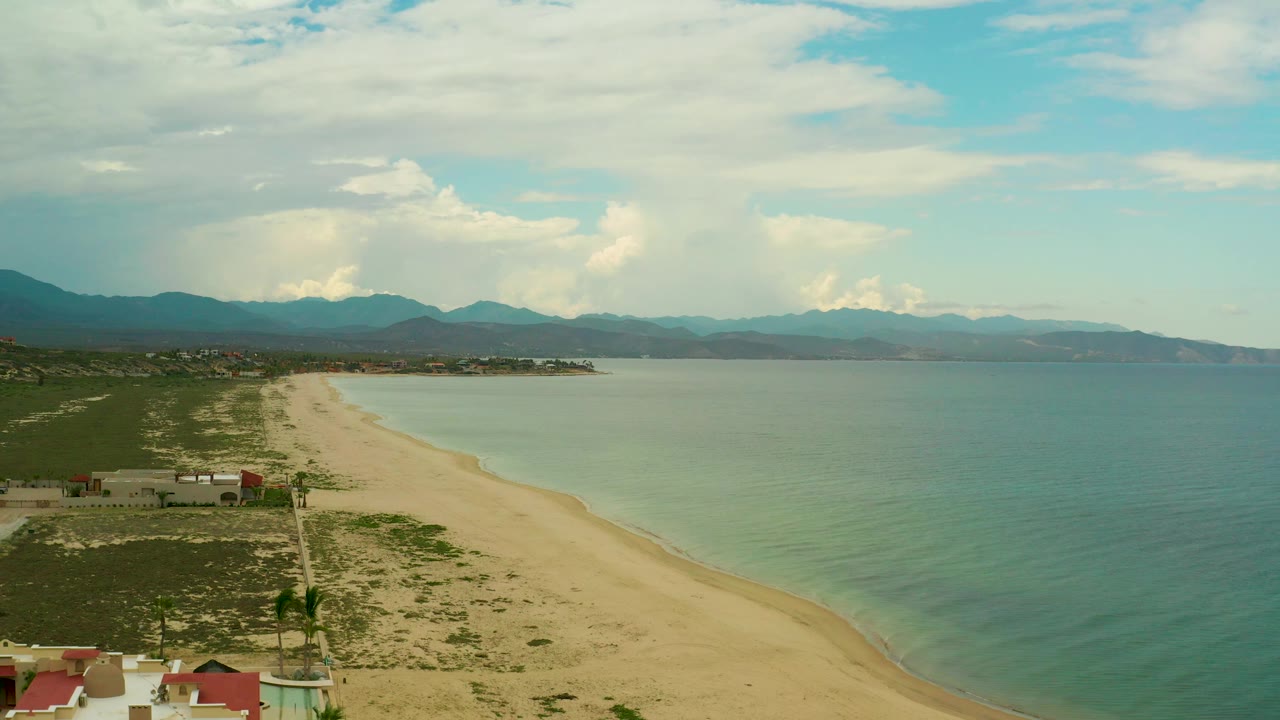 Aerial beach and sea view with green scrubland, shoreline structures, and cloudy mountain backdrop.