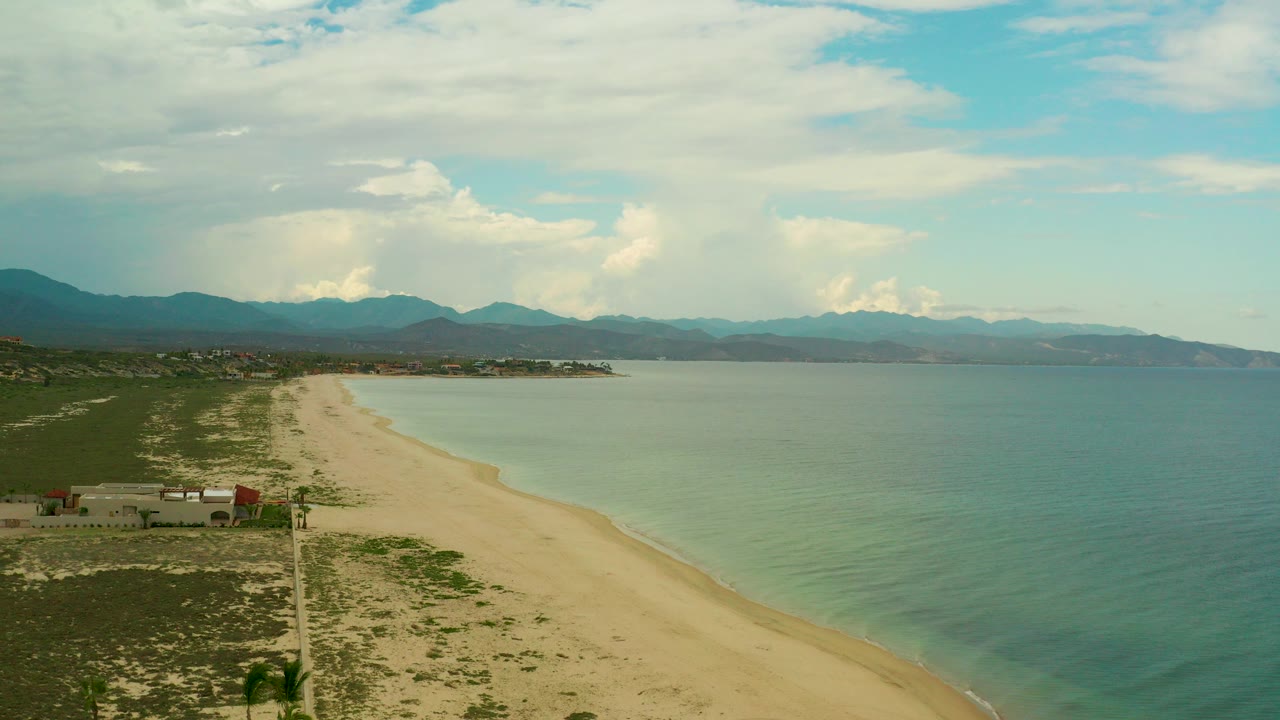 Aerial frame over La Ribera, Baja California Sur, with pale sand curving along turquoise-green water, distant mountains, cloud cover, and no visible people.