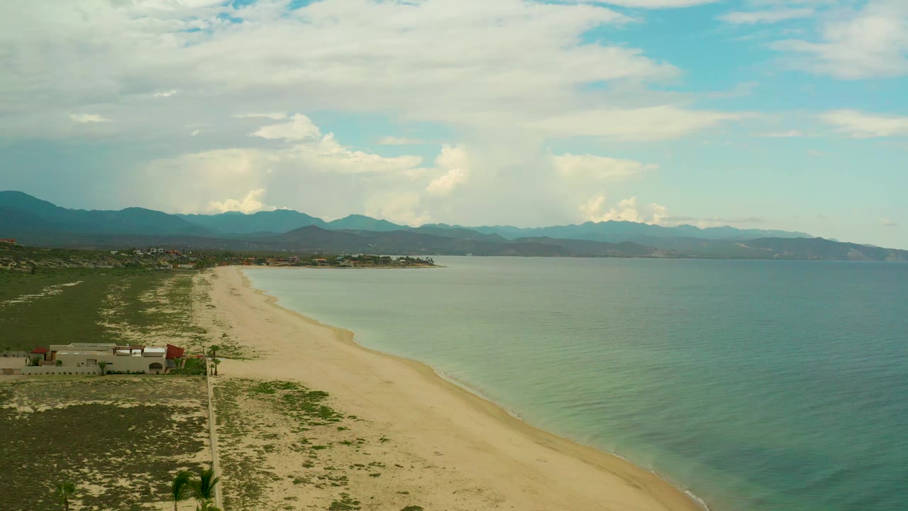 Aerial view in La Ribera, Baja California Sur, showing the beach running diagonally beside calm water, small shoreline buildings, distant mountains, and no visible people.