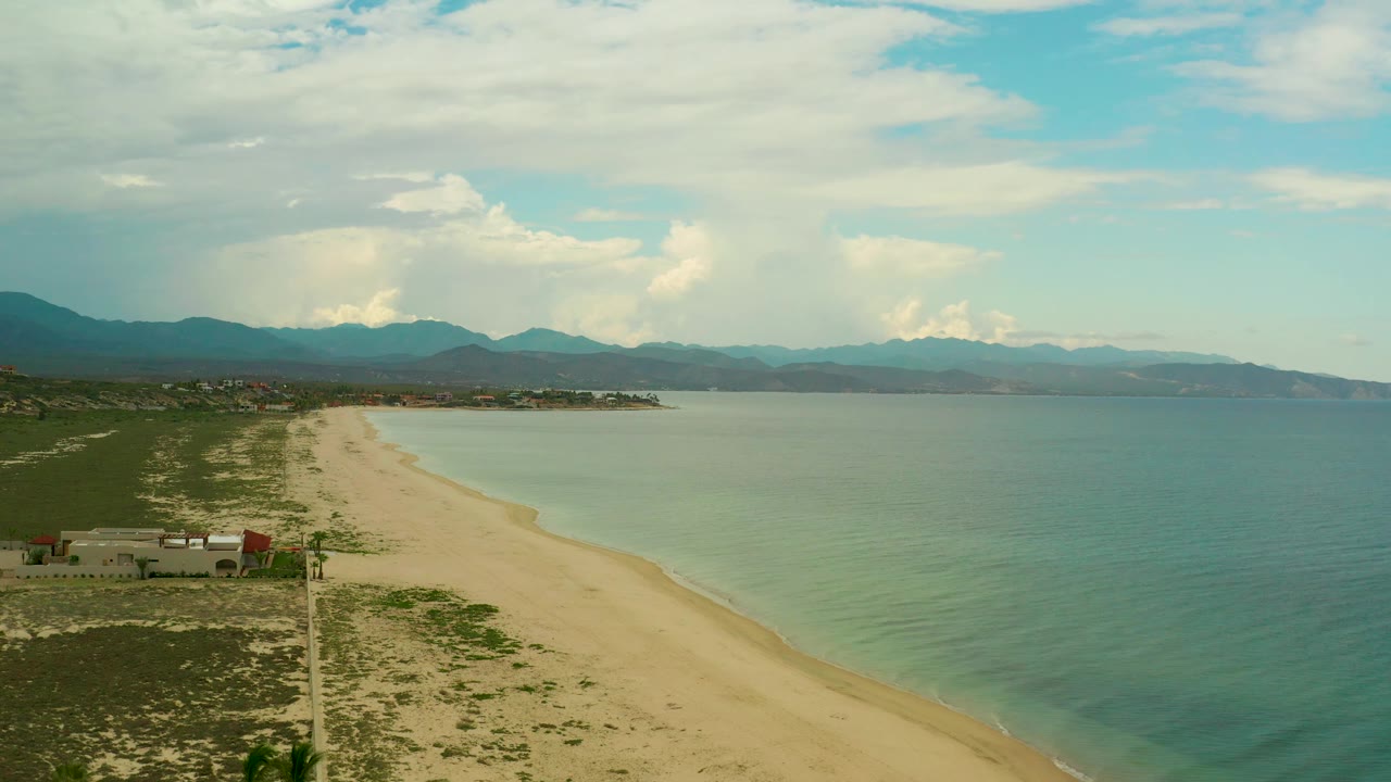 The La Ribera coastline appears from above with a long sand strip, still sea surface, low brush, a few coastal structures, mountains on the horizon, and no visible people.