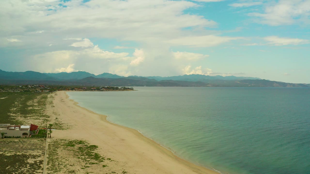 Aerial frame from La Ribera, Baja California Sur showing a sandy shoreline, clear coastal water, small structures at lower left, distant mountains, clouded sky, and no visible people.