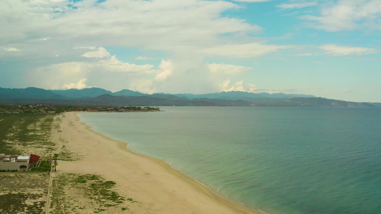 La Ribera beach fills the left side of the frame while blue-green water spreads right, with a few small buildings near the shore, layered mountains in back, and no visible people.