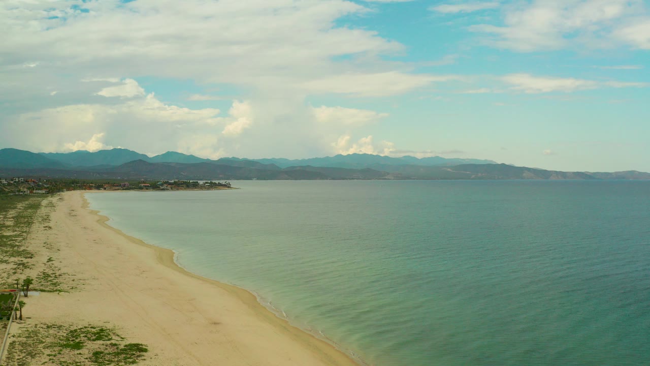 Opening aerial frame over La Ribera, Baja California Sur, showing pale sand curving along blue-green water, distant mountains, white clouds, and no visible people.