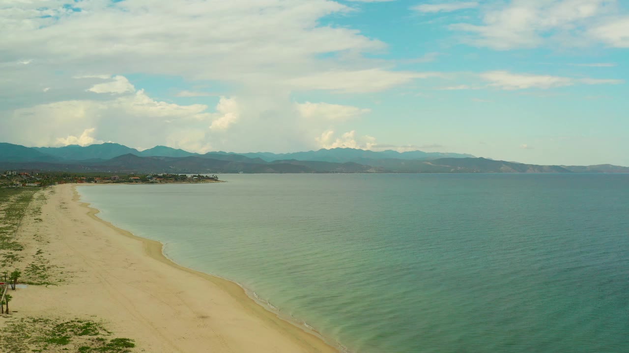 At 0:02, the aerial view shows the beach curve more clearly against shallow surf, with open sea, distant Baja California Sur mountains, white clouds, and no visible people.