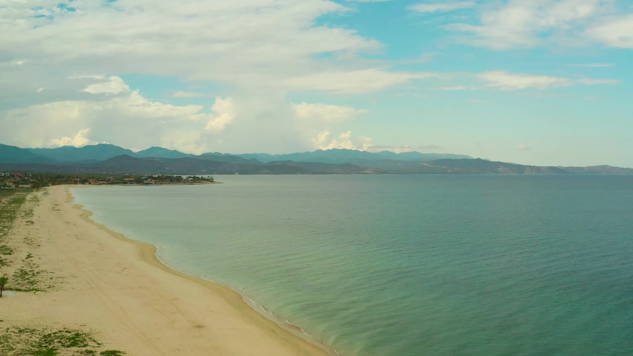 At 0:03, La Ribera's sand runs diagonally from the lower left toward the midframe, with calm sea to the right, hazy mountains beyond, and no visible people.