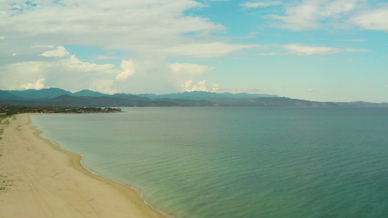 Opening aerial frame over La Ribera, Baja California Sur, with pale sand at left, green-blue water at right, distant mountains on the horizon, scattered clouds, and no visible people.