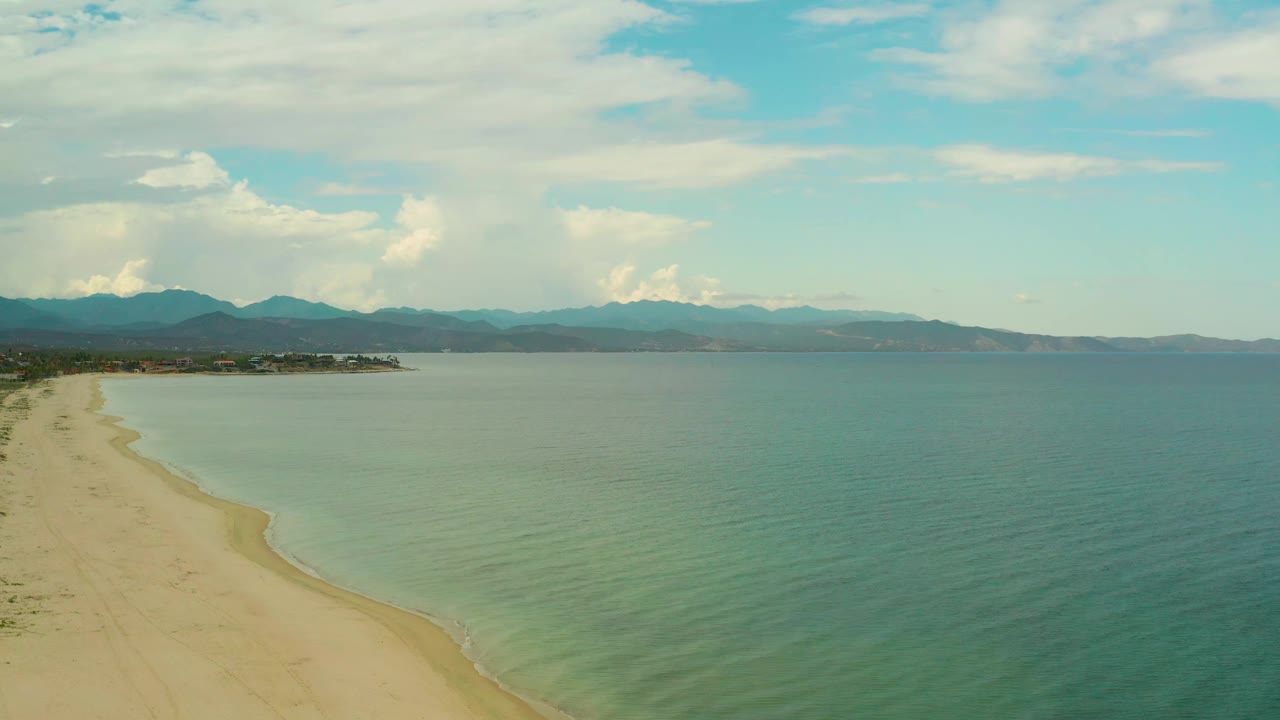 Aerial frame in La Ribera, Baja California Sur, with the shoreline curving down the left side, mild wave bands near shore, hazy mountain ridges across the background, and no visible people.