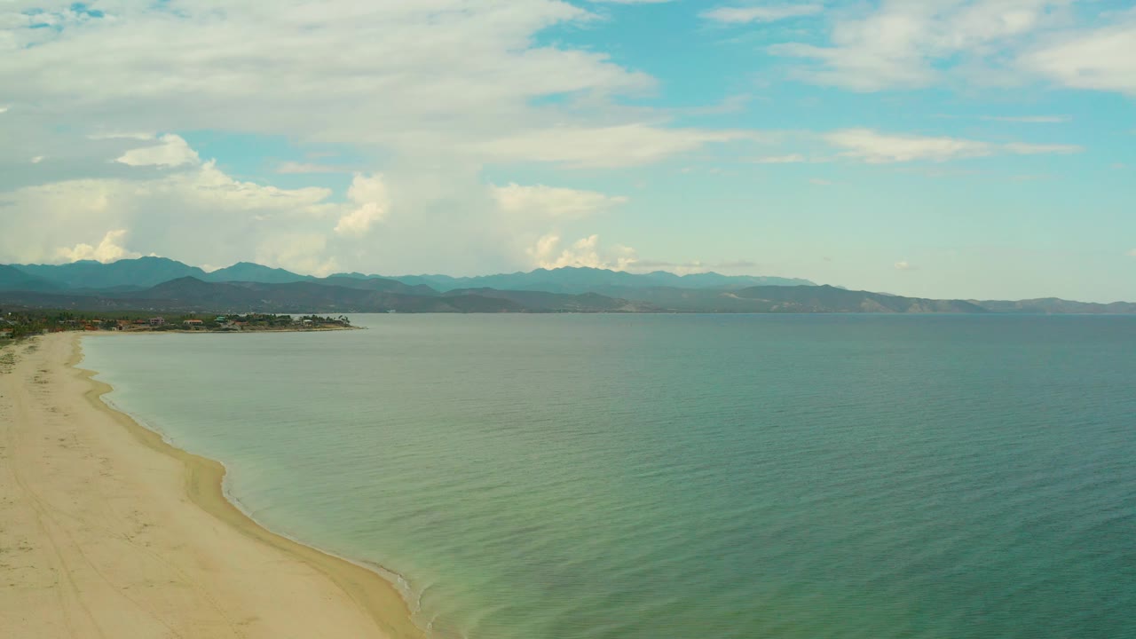 Mid-clip aerial frame over La Ribera, Baja California Sur, with pale beach visible at lower left, calm water extending to the horizon, mountain silhouettes in back, soft clouds, and no visible people.