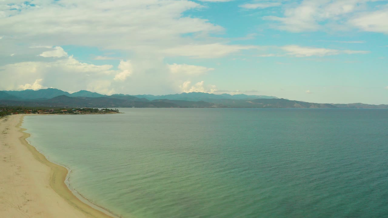 Aerial view over La Ribera, Baja California Sur with pale sand at left, blue-green water across the frame, distant mountains, bright clouds, and no visible people.
