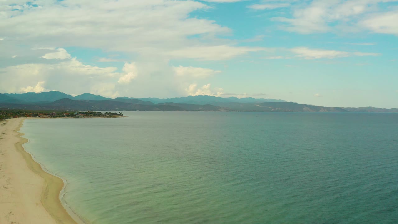 Midway through the clip, the aerial view shows shallow water near the sand, a narrow beach strip at left, low mountains beyond the bay, bright cloud formations, and no visible people.
