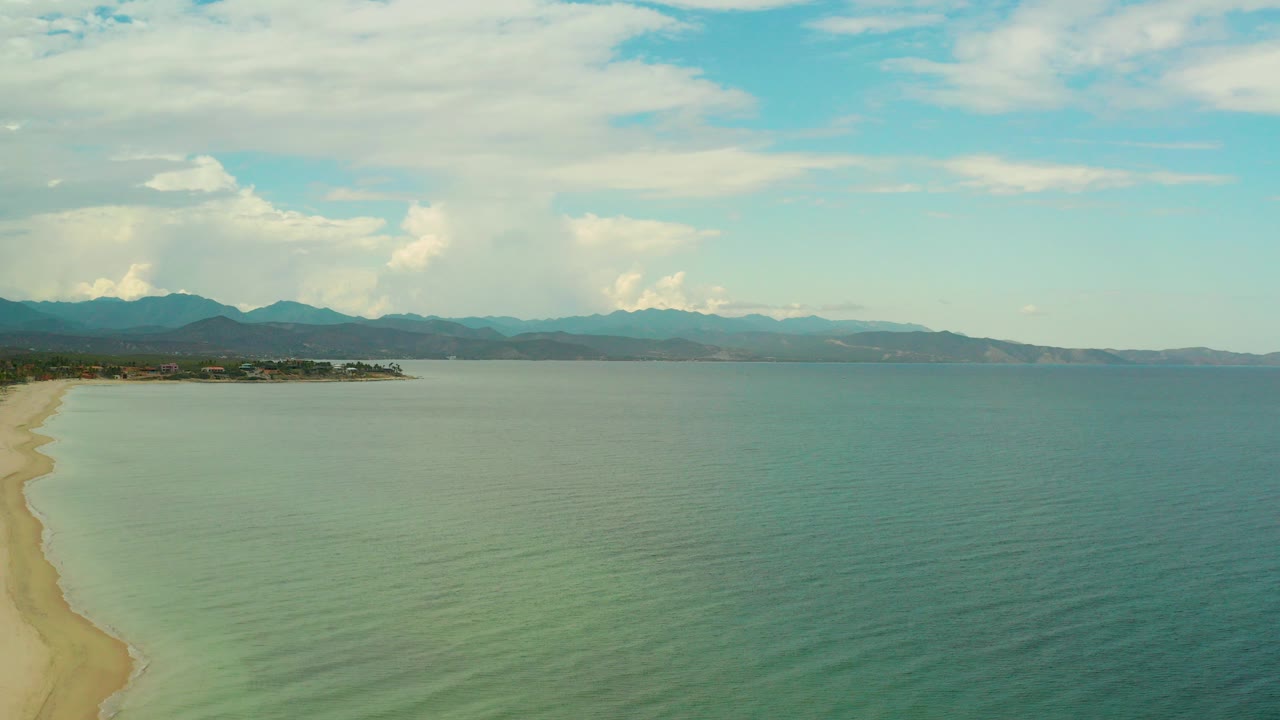Opening aerial frame over La Ribera, Baja California Sur, showing a curving sand beach at left, turquoise-green water in front, distant mountains, clouded blue sky, and no visible people.