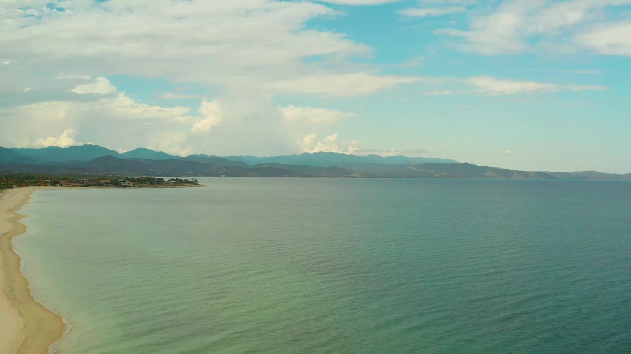 Aerial view at 0:01 in La Ribera with the pale beach still tight to the left edge, broad green-blue water filling the frame, mountains along the horizon, bright clouds, and no visible people.