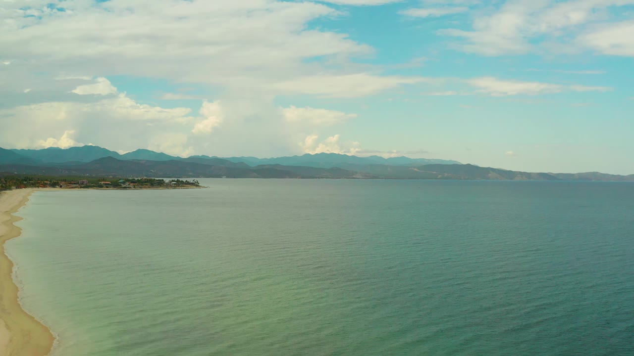 Aerial frame over La Ribera at 0:02 showing the sandy shoreline running down the left side, open water across most of the image, layered mountains in back, high clouds, and no visible people.