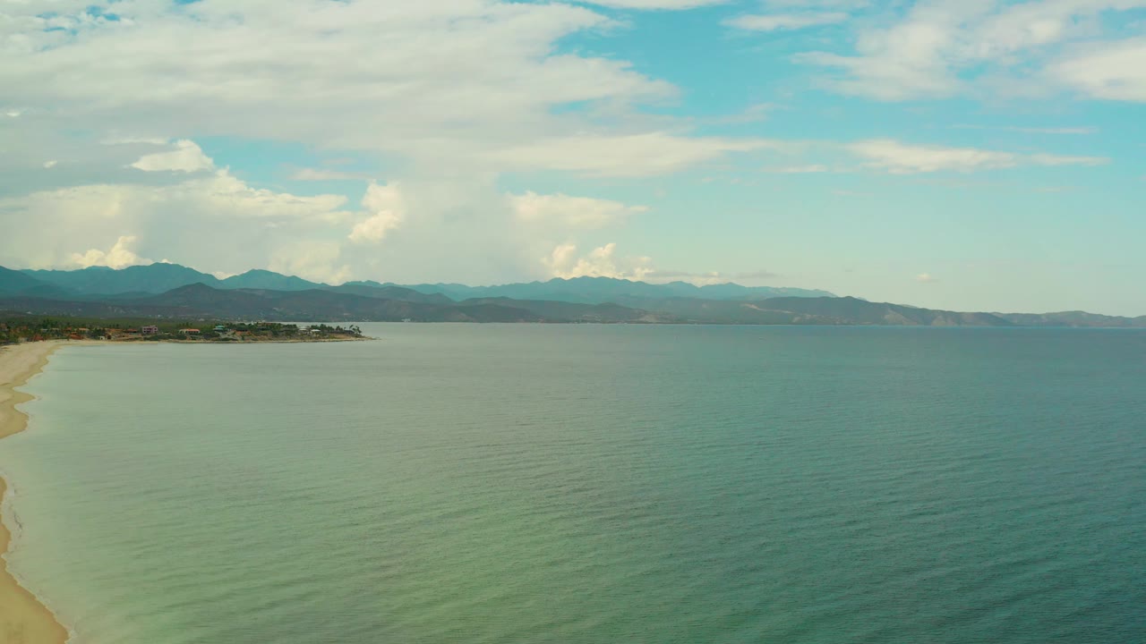Mid-clip aerial frame in La Ribera with the beach edge partly cropped at left, calm coastal water in the foreground, low land in the distance, mountains beyond, clouded sky, and no visible people.