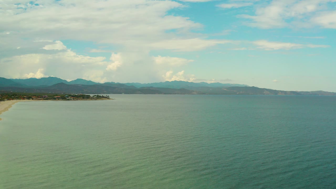 Opening aerial frame over La Ribera, Baja California Sur, with green-blue ocean in the foreground, sandy beach at left, distant mountains on the horizon, and no visible people.