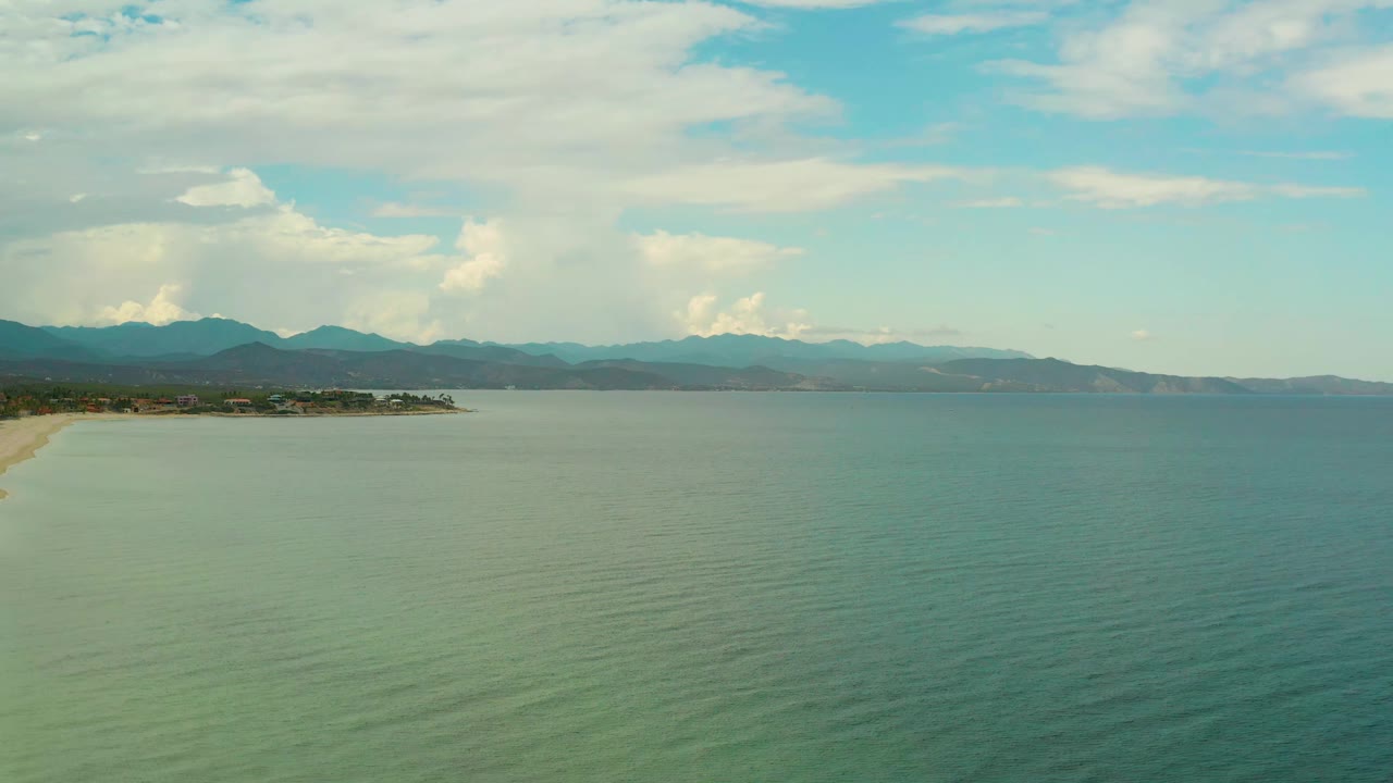 Aerial view in La Ribera, Baja California Sur, showing a broad sheet of ocean water, a narrow beach edge on the left, layered mountains in the distance, and no visible people.