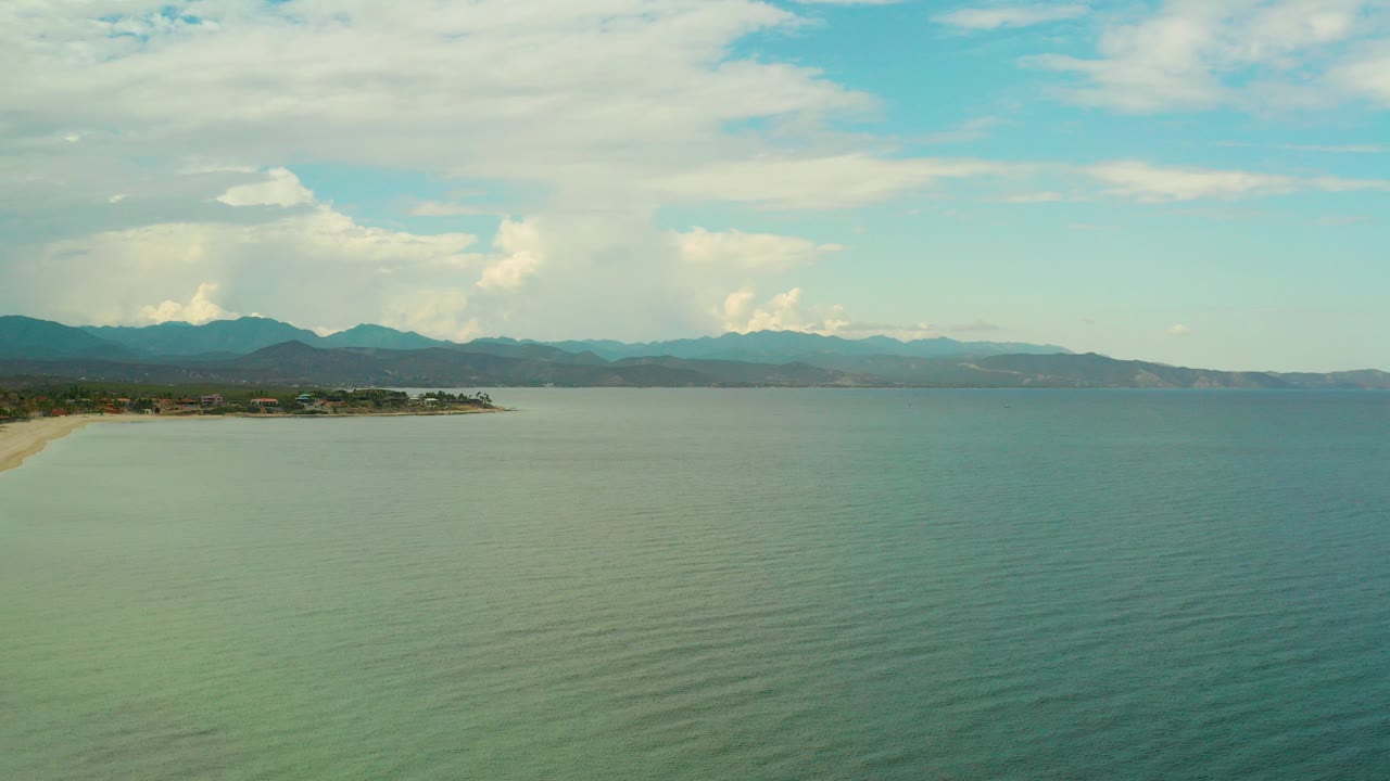 Wide aerial frame over La Ribera, Mexico, showing rippled blue-green sea, a pale beach edge on the left, tree-lined shore, distant mountains, broad clouds, and no visible people.
