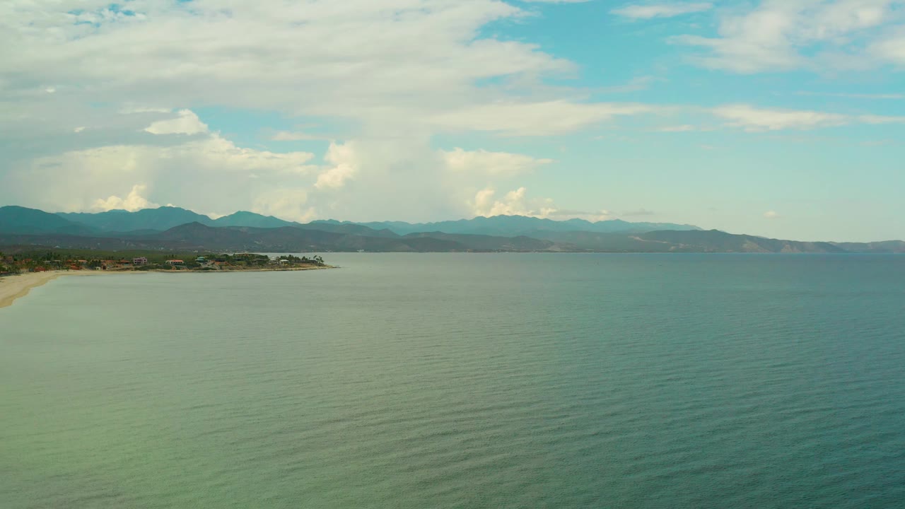 La Ribera, Mexico coastline from above, with open rippled sea, a thin sand line at left, dark green shoreline trees, hazy mountain ridges, high clouds, and no visible people.