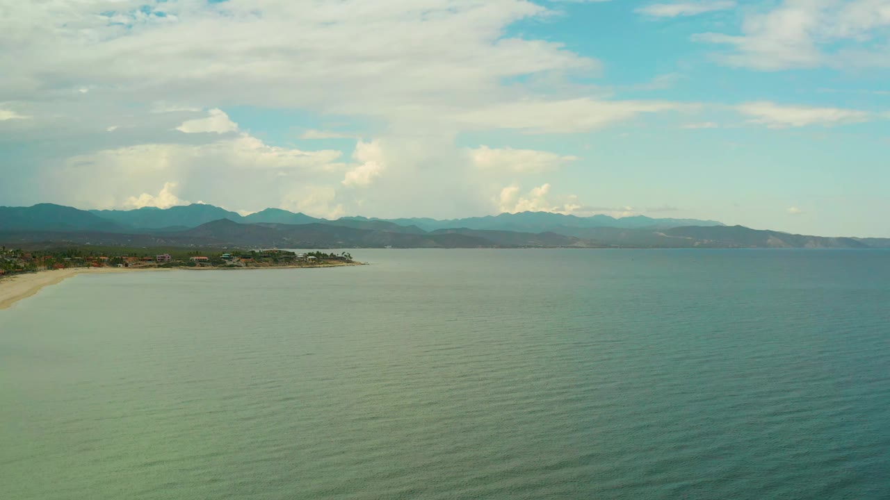 Aerial coastal view with open water, shoreline, distant buildings, mountains, and clouds.
