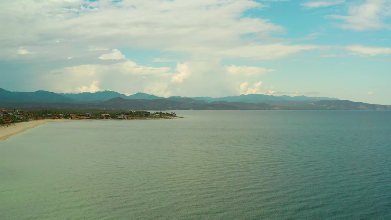 Opening frame of La Ribera, Baja California Sur with blue-green ocean in front, a sandy coast at left, distant mountains, high clouds, and no visible people.