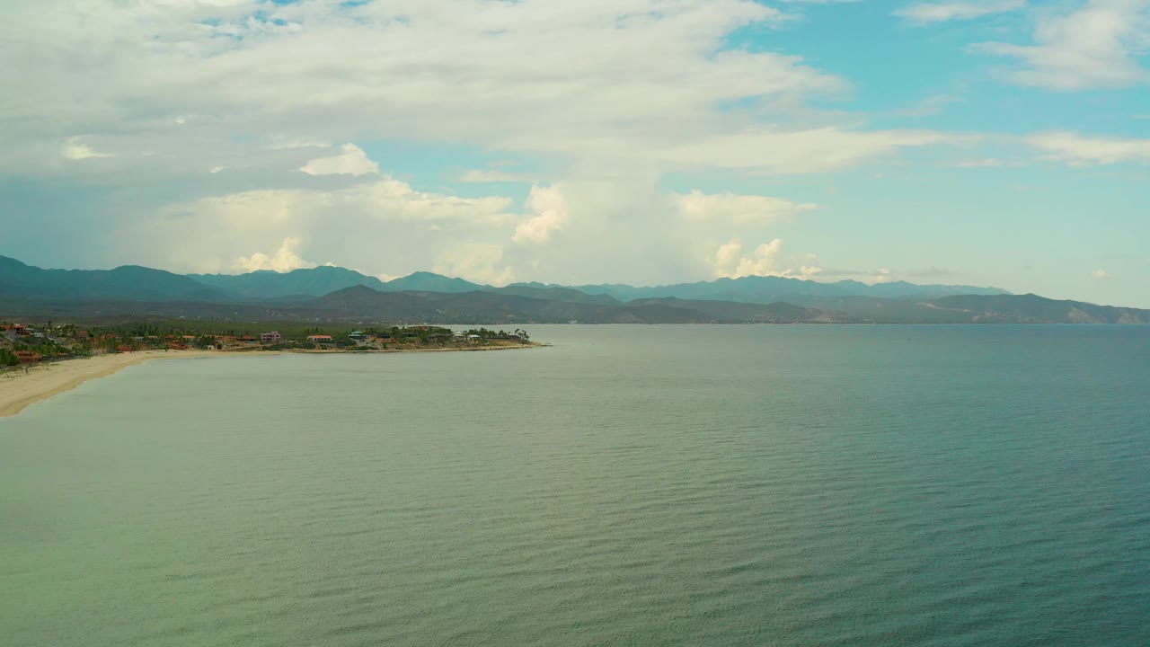 Wide La Ribera ocean view with faint water ripples, a curved sandy beach along the left edge, layered mountains on the horizon, and no visible people.