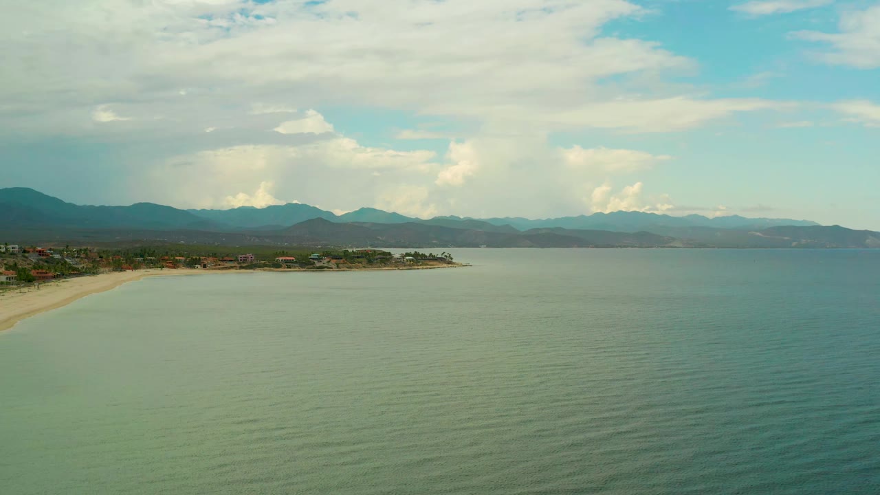 Wide aerial coastline with calm ocean, beach on the left, and mountain ridges beyond.