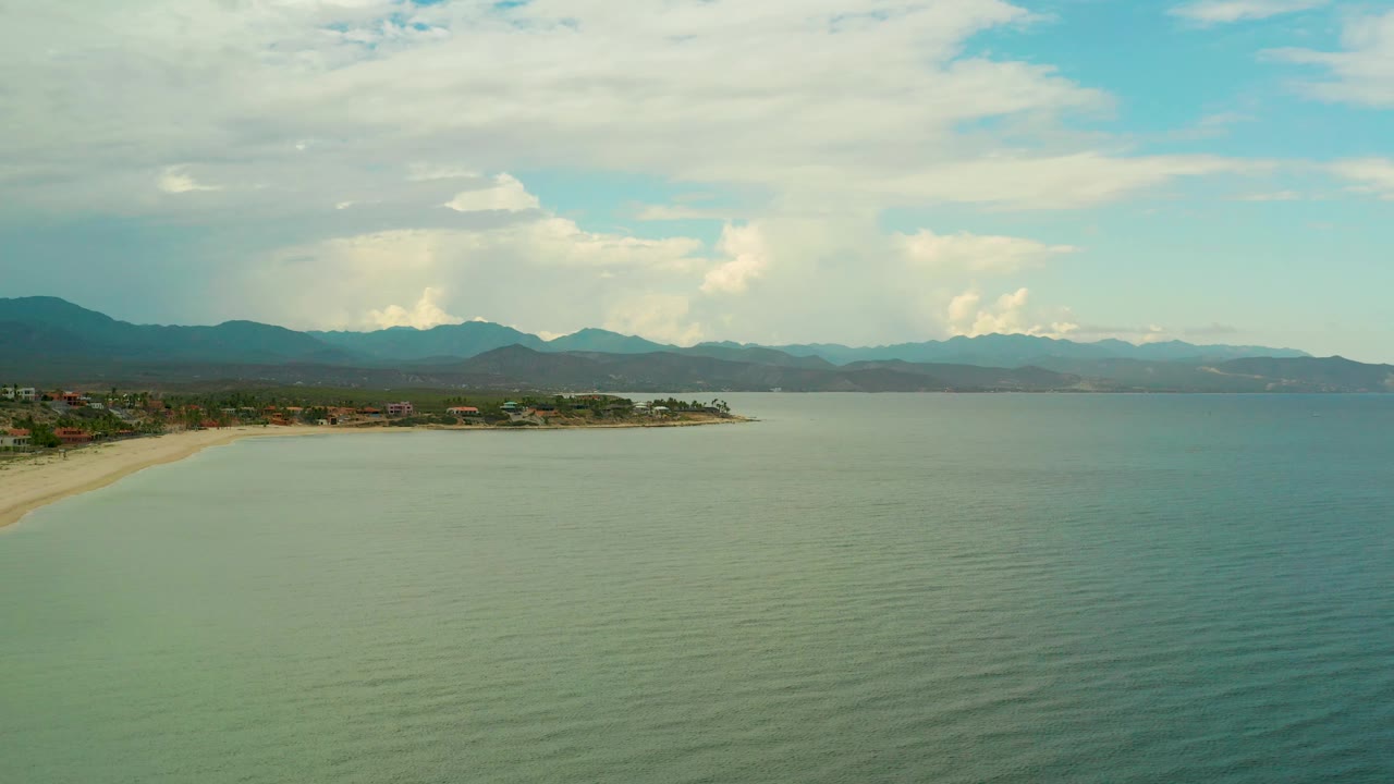 La Ribera aerial frame with the shoreline still anchored on the left edge, broad green water in front, distant dark hills and white cloud towers behind, and no visible people.