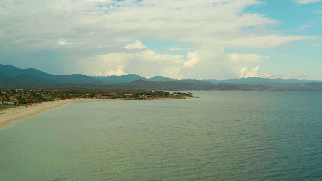 A high aerial view over La Ribera, Baja California Sur shows calm green-blue sea, a sandy shoreline at left, distant mountains, bright clouds, and no visible people.