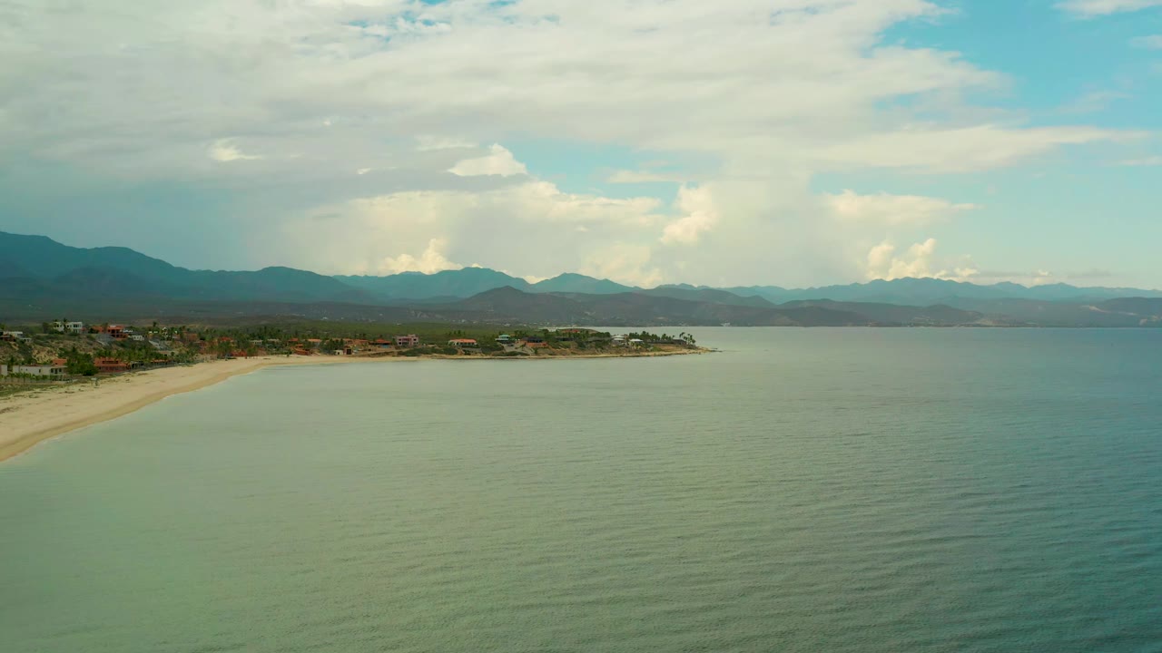 The La Ribera coast remains in a wide aerial frame, with shallow-looking water across the foreground, beach and small buildings to the left, mountains on the horizon, and no visible people.