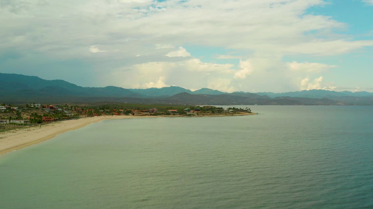 Wide aerial view over La Ribera, Baja California Sur, with green-blue ocean in front, a pale beach curving left, coastal buildings along shore, distant mountains, cloud cover, and no visible people.