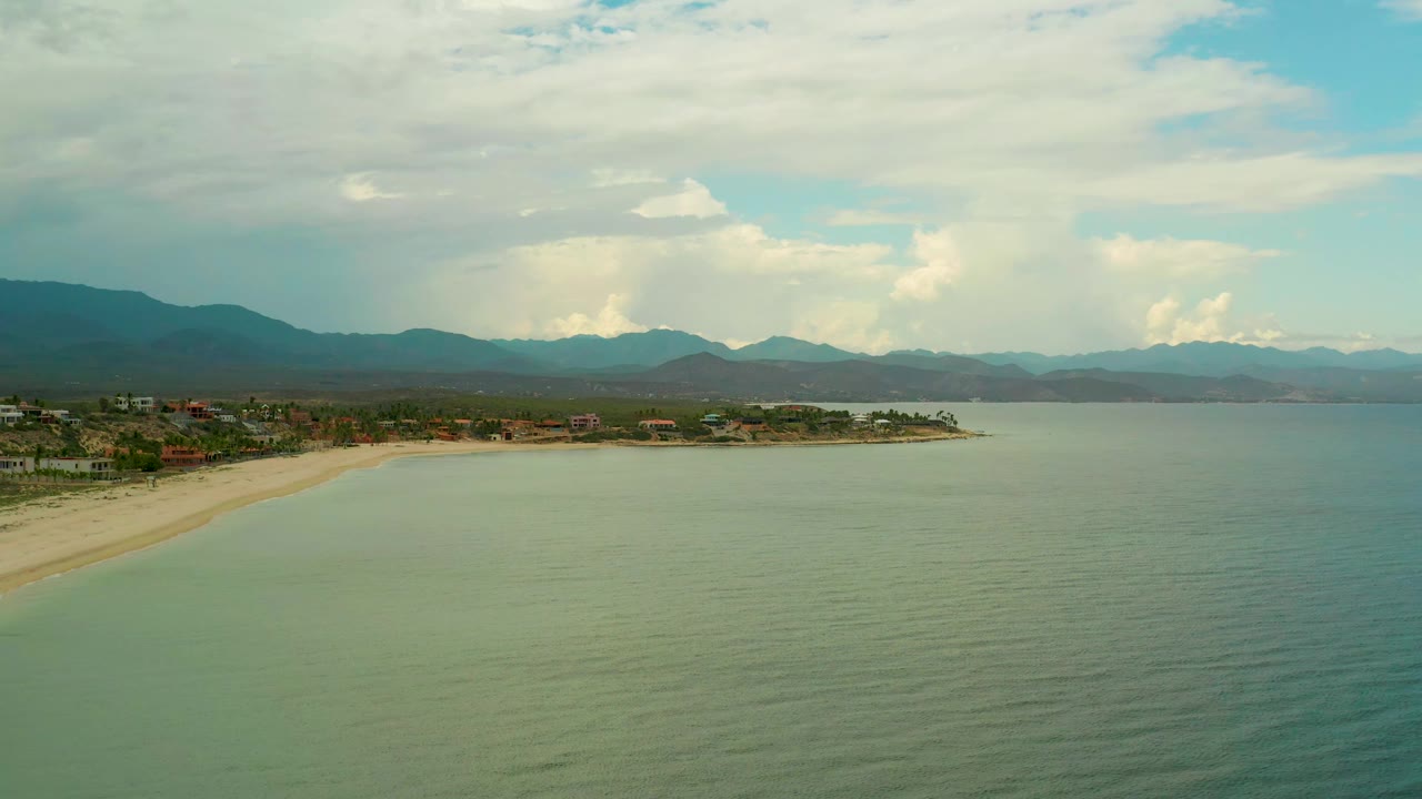 Aerial frame over La Ribera coastline showing calm green-blue water across the foreground, a sandy shoreline at left, low coastal buildings, hazy mountain ridges, thick clouds, and no visible people.