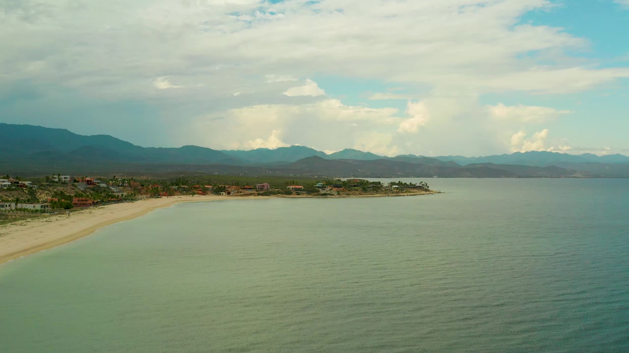 La Ribera beach and bay fill the frame, with smooth water texture in the foreground, a narrow strip of sand, clustered coastal buildings, distant mountains, bright cloud patches, and no visible people.