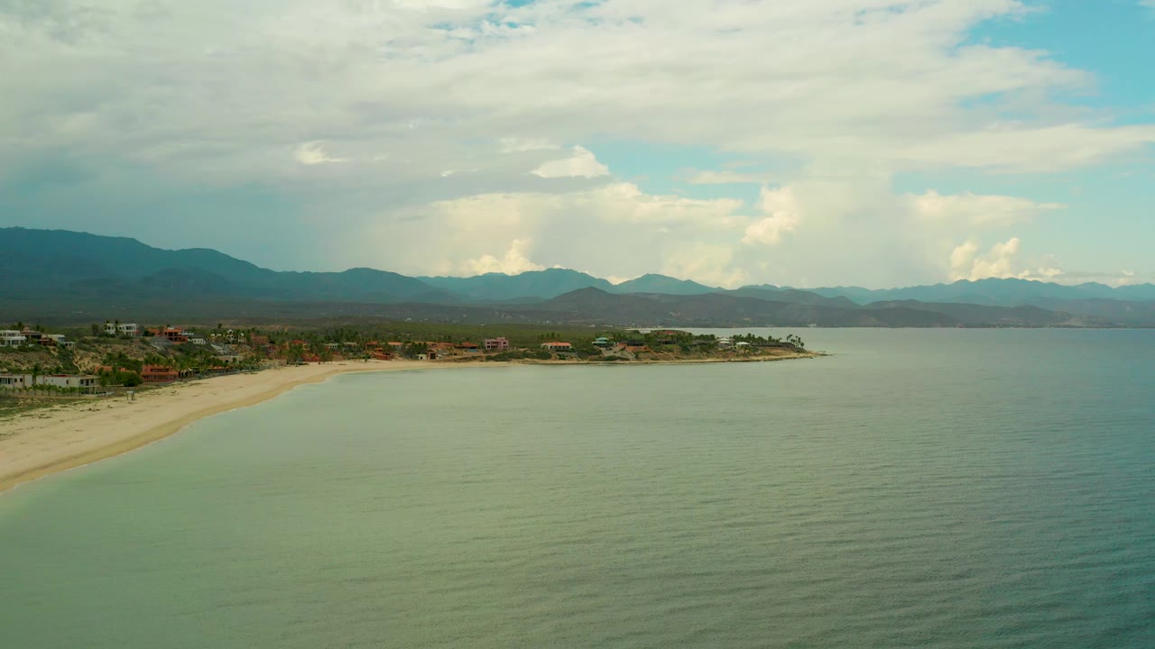 Curving sandy beach beside green-blue ocean with mountains under clouds.