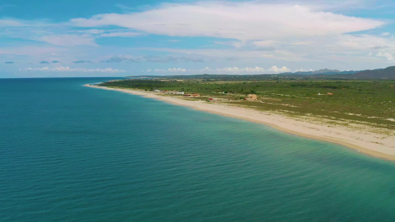 Aerial view over La Ribera, Baja California Sur, with blue-green sea in the foreground, a diagonal sandy beach, low green vegetation inland, distant hills, and no visible people.